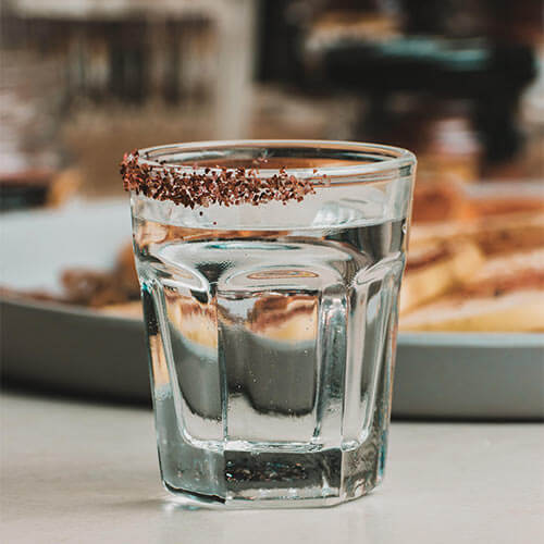 Vaso de agua con borde decorado con sal y chocolate rallado, sobre una mesa. Fondo desenfocado.