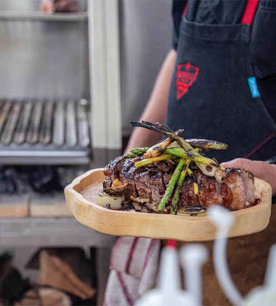 Persona sosteniendo un plato de carne a la parrilla con espárragos, listo para servir en un entorno de barbacoa.