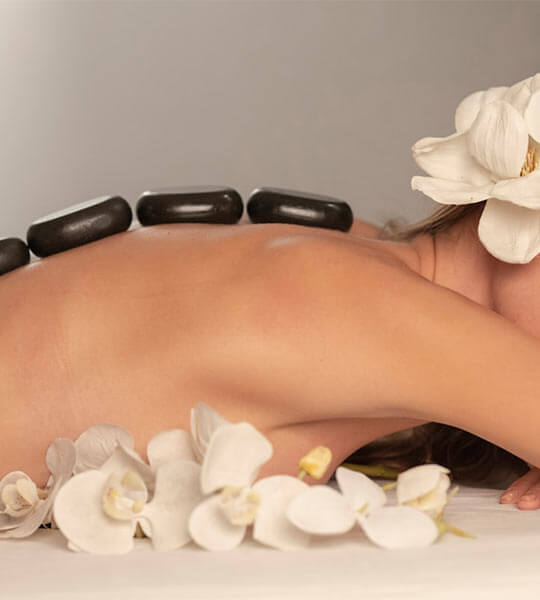Mujer recostada boca abajo con piedras calientes en la espalda y flores blancas decorativas alrededor.