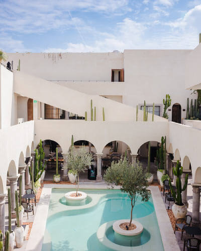 Patio con piscina, rodeada de arcos y cactus, en un edificio blanco iluminado por el sol.