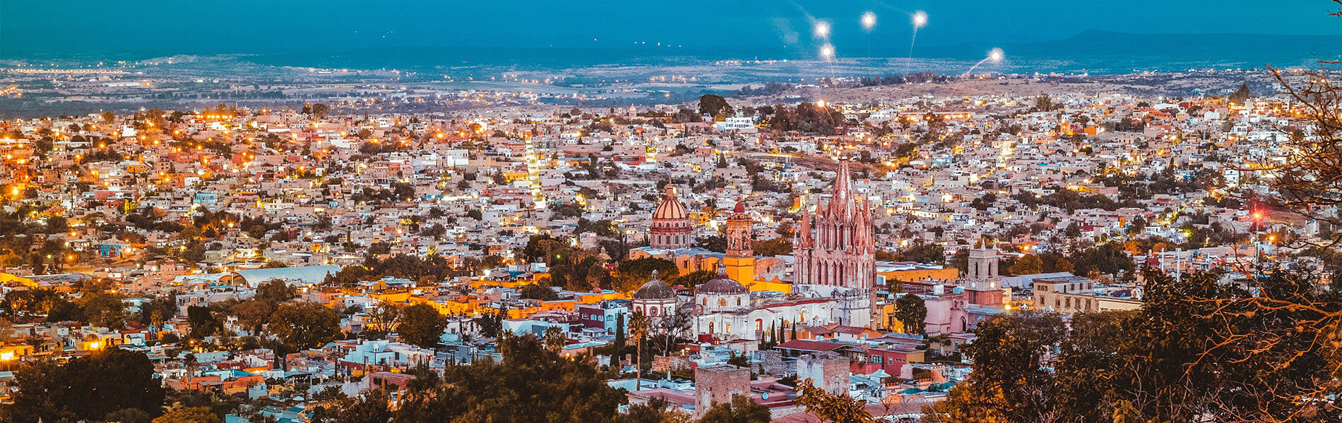 Vista panorámica nocturna de una ciudad iluminada con arquitectura colonial, rodeada de colinas y un cielo azul oscuro.