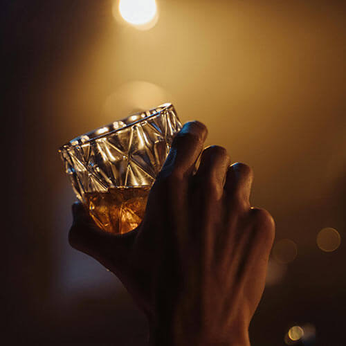 Mano sosteniendo un vaso de whisky contra una luz cálida y difusa en el fondo.