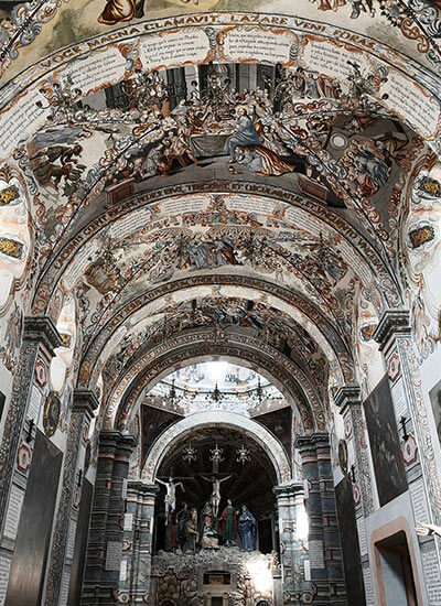 Interior de una iglesia con techo pintado de escenas religiosas y un altar detallado al fondo.