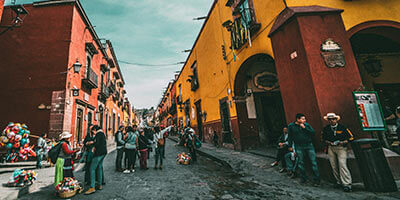 Calle colorida con edificios coloniales, personas caminando, y vendedores de globos, ambientada en un día soleado.