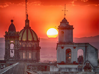 Cúpulas de una iglesia al atardecer con el sol poniéndose en el horizonte y un cielo rojo intenso.