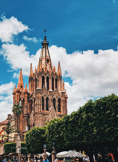 Iglesia con torres góticas adornadas contra un cielo azul y un primer plano de árboles verdes y personas caminando.