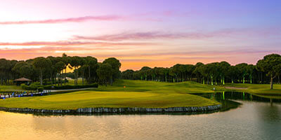 Campo de golf al atardecer, con césped verde y un lago, rodeado de árboles y un cielo colorido.
