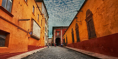 Calle de adoquines bordeada por edificios coloniales de tonos cálidos bajo un cielo parcialmente nublado.