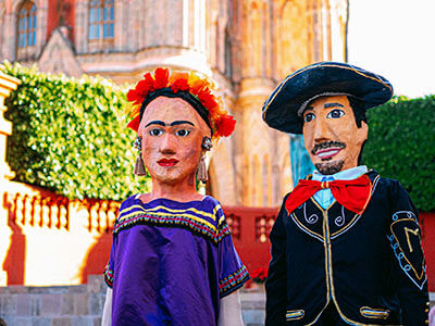 Dos mojigangas tradicionales mexicanas con atuendos coloridos en una plaza, con un edificio de fondo.