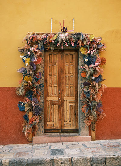 Puerta de madera decorada con abundantes adornos secos de colores en una pared amarilla y roja.