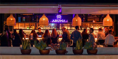 Personas en un bar llamado Aruma, con lámparas colgantes y plantas decorativas al frente.