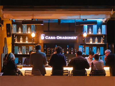 Personas frente a un bar iluminado con botellas y el letrero "Casa Dragones" al fondo.
