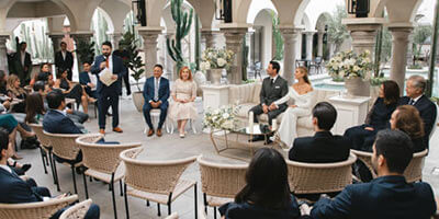 Grupo de personas sentadas en un evento formal al aire libre, con pilares y plantas decorativas alrededor.