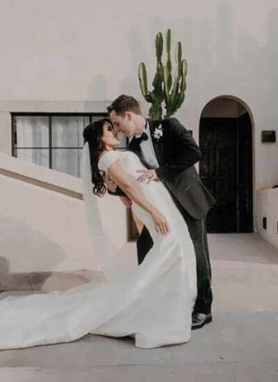 Pareja de novios besándose, él con traje negro y ella con vestido blanco; de fondo, un cactus y un edificio beige.