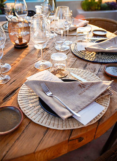 Mesa de madera al aire libre con manteles individuales, copas y cubiertos dispuestos para una cena elegante.