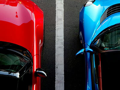 Dos autos, uno rojo y otro azul, estacionados paralelos con una línea blanca en el centro, vistos desde arriba.
