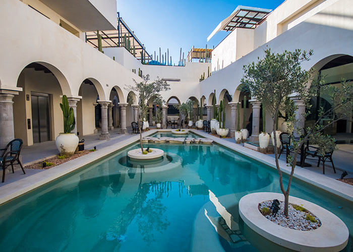 Patio con piscina rodeada de arcos, plantas en macetas blancas y sillas; cielo despejado arriba.
