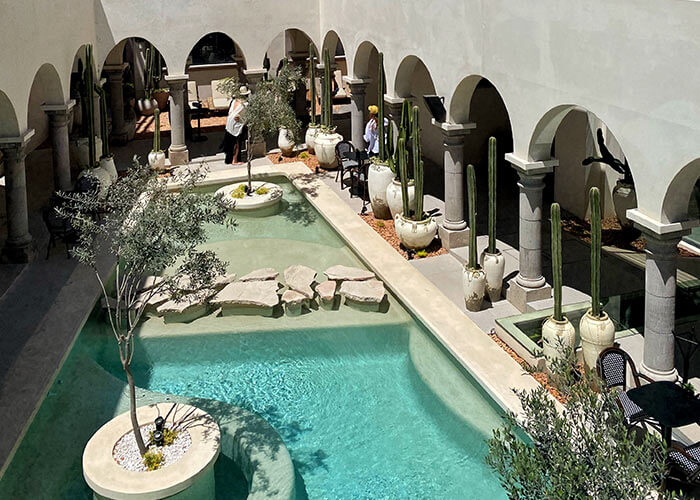 Patio con piscina, plantas en macetas y columnas arqueadas. Personas de pie junto a las paredes. Ambiente cálido y relajante.