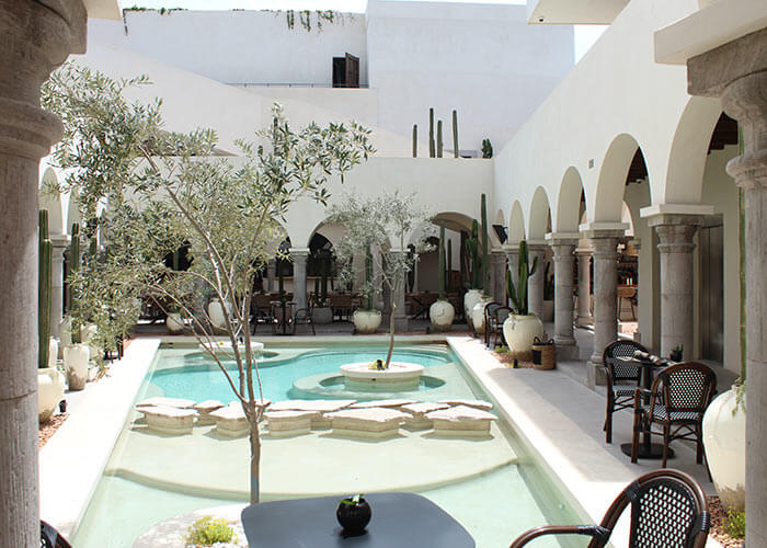 Patio con piscina central rodeada de columnas, plantas y sillas, en un entorno luminoso de estilo mediterráneo.