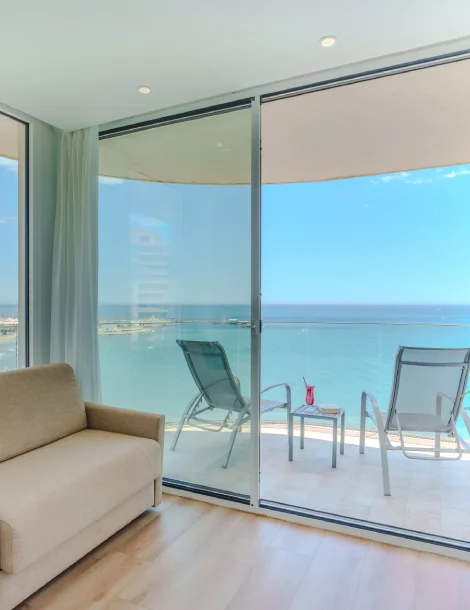 Vista al mar desde sala con sofás y balcón con sillas y bebida.