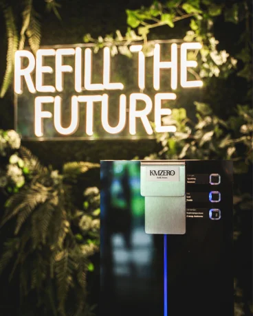 Máquina expendedora con letrero "Refill the Future" y vegetación de fondo.