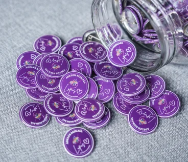 Botones morados con inscripciones y dibujos, esparcidos desde un frasco de vidrio sobre tela gris.