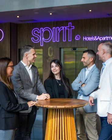 Cinco personas charlan alrededor de una mesa, con un cartel de "spirit Hotels & Apartments" detrás.