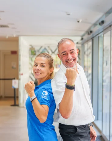 Dos personas sonriendo y posando de espaldas en un pasillo iluminado.