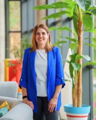 Mujer de pie junto a ventana con plantas, vestida con blazer azul y camisa blanca.