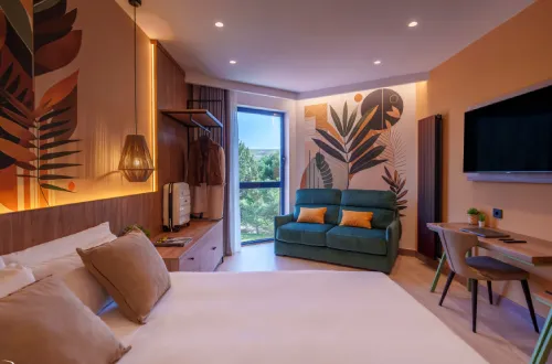 Habitación moderna con cama, sofá, televisor, y decoración de hojas; ventana al paisaje exterior.