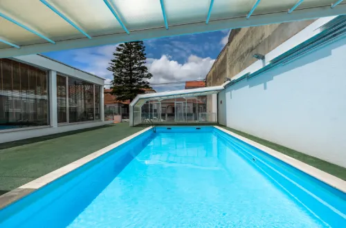 Piscina al aire libre con techo de vidrio y paredes blancas bajo un cielo azul y nublado.