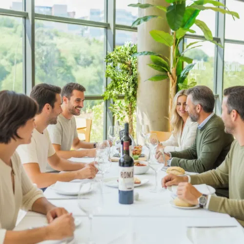 Personas cenando felices en un restaurante con vista a un paisaje verde y una botella de vino sobre la mesa.