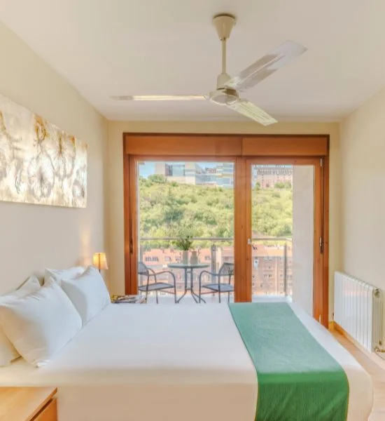 Habitación luminosa con cama, vista a la ciudad y balcón con mesa y sillas.