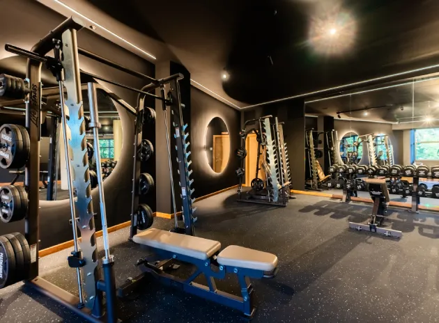 Gimnasio moderno con máquinas de pesas, espejos y bancos sobre un suelo oscuro.