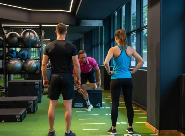 Personas entrenando en un gimnasio, dos observan mientras una salta sobre una caja pliométrica.