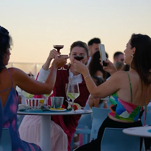 Tres personas brindan en una terraza al atardecer, mientras una de ellas toma una foto con el móvil.