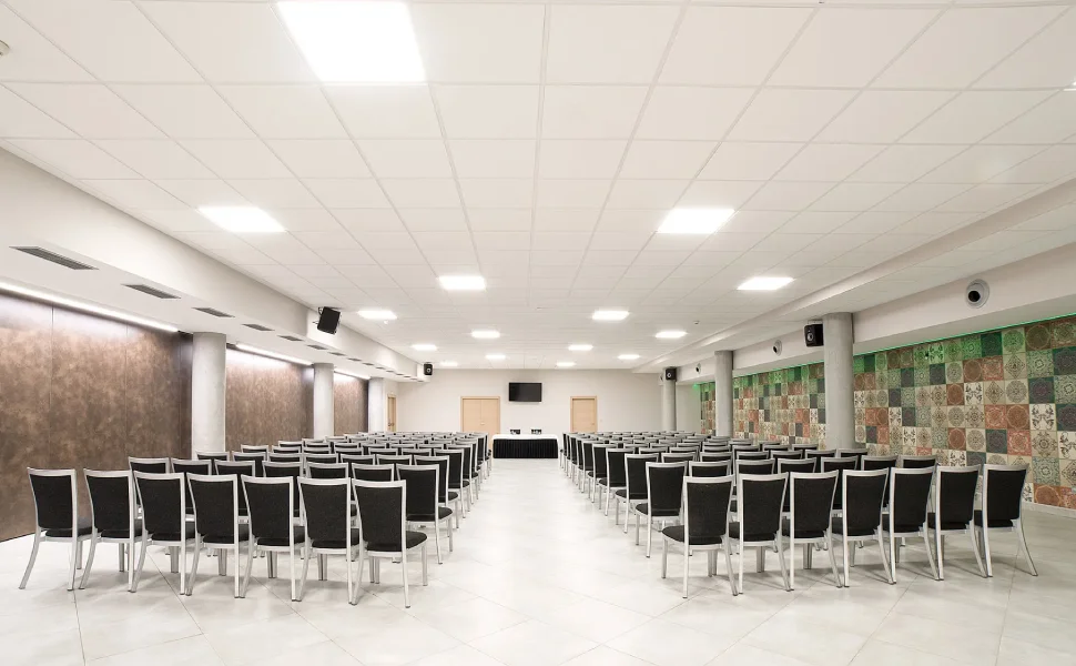 Sala de conferencias vacía con filas de sillas, pantalla y mesa al frente.