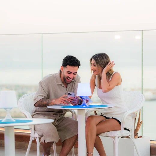Pareja riendo mientras miran un teléfono en una terraza con vista al mar.