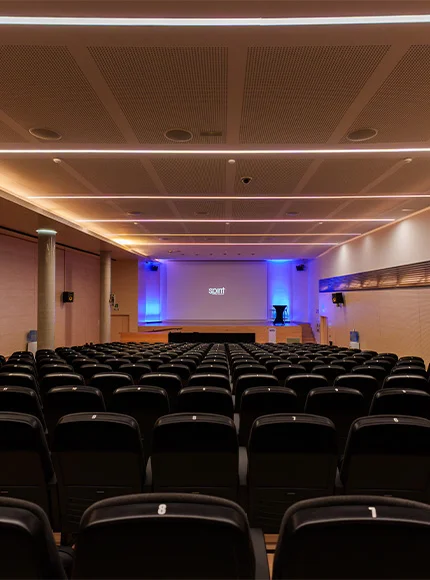 Auditorio vacío con filas de asientos, pantalla grande y luces azules en el frente.