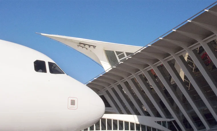 Nariz de un avión junto a una estructura arquitectónica moderna con cielo azul.