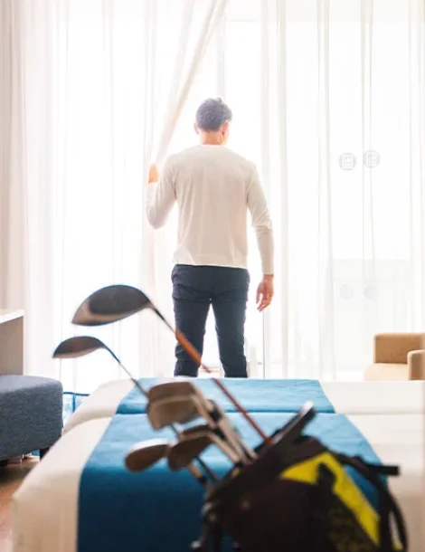 Hombre de pie junto a ventana con cortinas; palos de golf en primer plano.