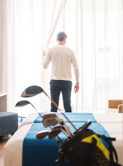 Hombre de pie junto a ventana con palos de golf en primer plano, en una habitación luminosa.