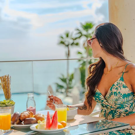Mujer comiendo en una terraza con vista al mar y palmeras, con desayuno servido en la mesa.