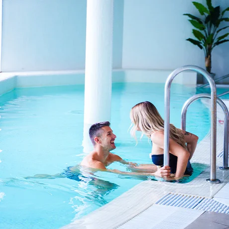 Pareja sonriente en una piscina interior, ella sentada en el borde y él en el agua.