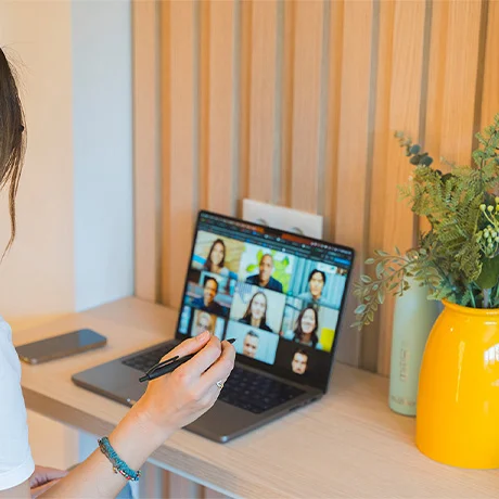 Mujer en videollamada desde una laptop en un escritorio con flores y un teléfono cercano.