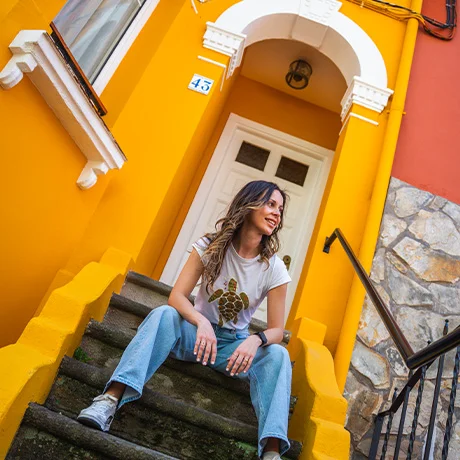 Mujer sentada en escaleras, usa camiseta blanca y jeans. Fondo de pared naranja con puerta blanca.