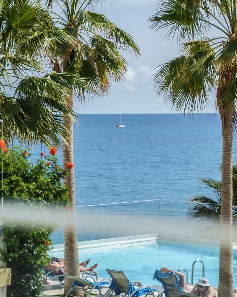 Piscina frente al mar con palmeras y personas relajándose en sillas, barco en el horizonte.