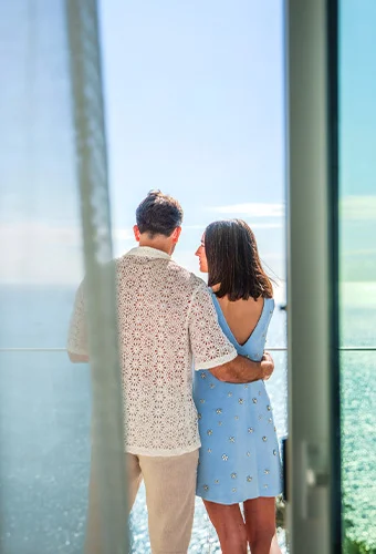 Pareja abrazada mirando el mar desde un balcón en un día soleado.