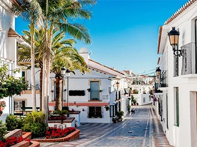 Calle de un pueblo mediterráneo con casas blancas, palmeras y cielo azul.