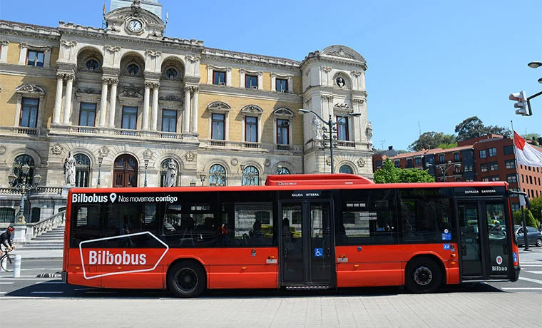 Autobús rojo de Bilbobus frente a un edificio histórico en un día soleado.